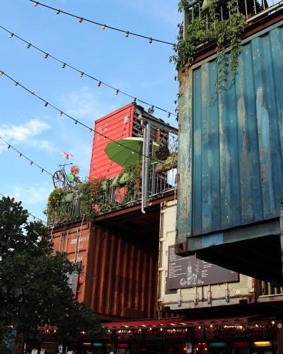 The image shows a multi-level outdoor structure constructed from stacked, repurposed shipping containers (blue, red, and brown), decorated with string lights, plants, and a bicycle on a balcony, under a blue sky.