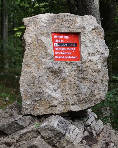A large, rough-hewn stone marker set in a forest clearing, featuring a red plaque with white text reading "Hinteri Egg 1169 m" and "Höchster Punkt des Kantons Basel-Landschaft".
