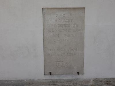 A close-up photograph of a rectangular stone memorial plaque mounted on a plain, light-colored exterior wall, featuring Latin text and a small carved emblem at the bottom.