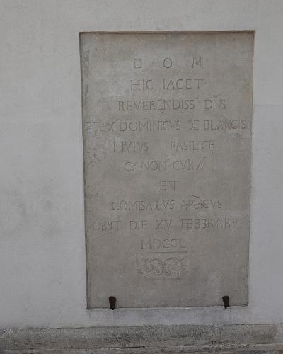 A close-up photograph of a rectangular stone memorial plaque mounted on a plain, light-colored exterior wall, featuring Latin text and a small carved emblem at the bottom.
