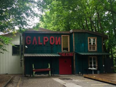 A teal wooden building with large red letters spelling "GALPON" and smaller red letters spelling "THEATRE" on the facade, surrounded by trees.