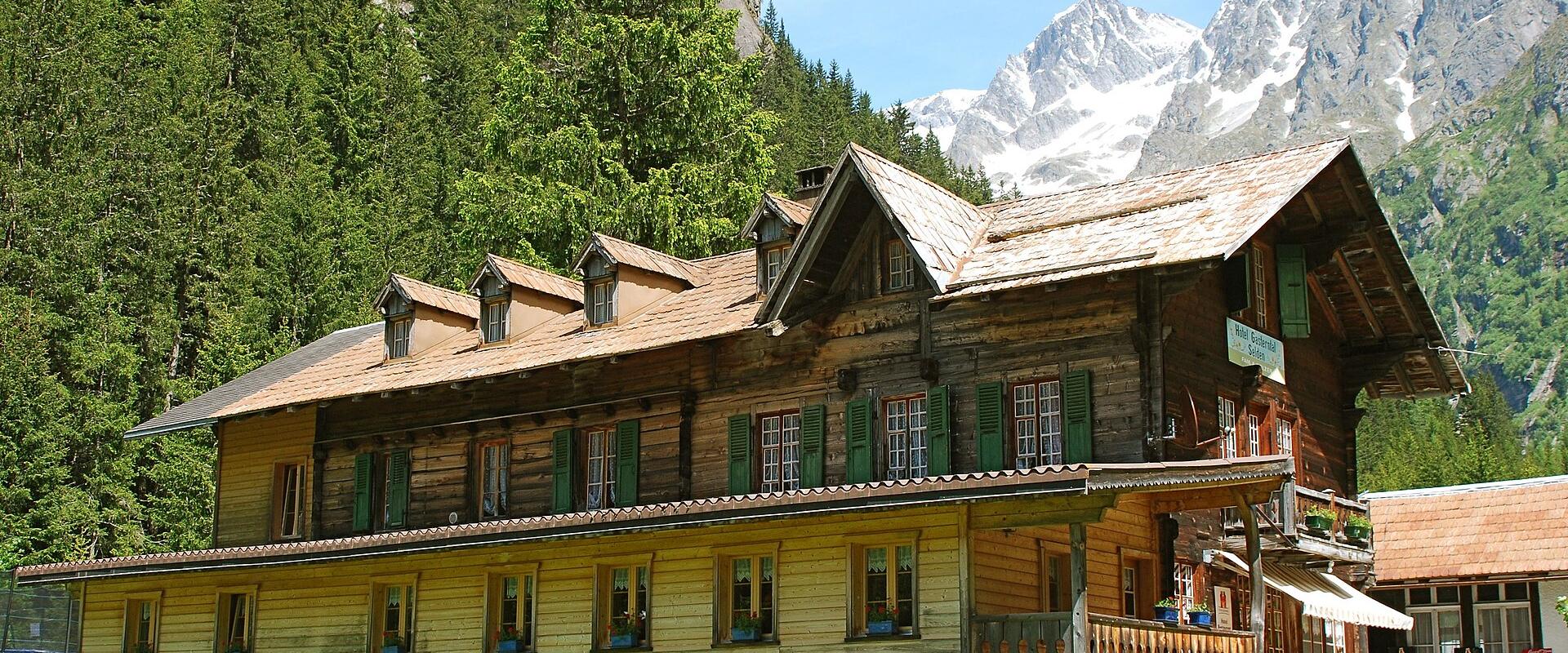 A large, traditional wooden chalet with green shutters and a stone foundation, set against a backdrop of steep, forested mountains and snow-capped peaks.