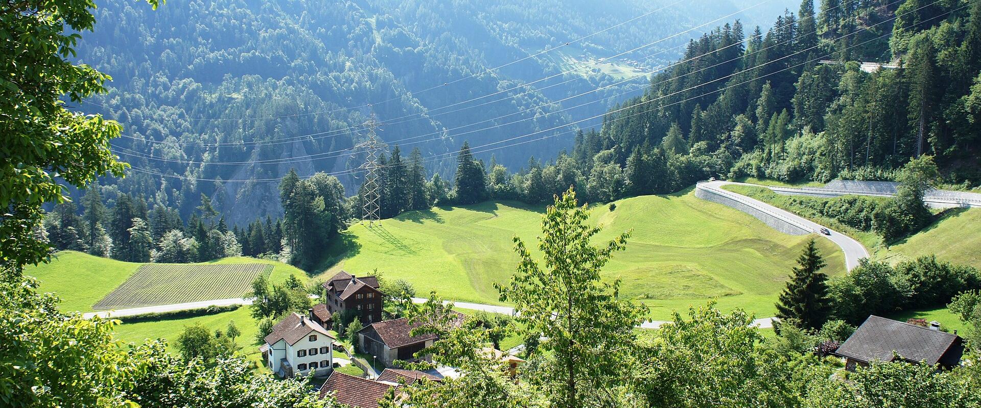 A scenic view of a small cluster of traditional houses and farm buildings nestled in a green alpine valley, with a winding road, forested slopes, and high-voltage power lines visible in the background.