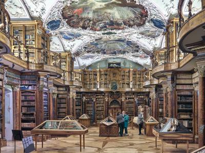 The image shows the interior of a grand, two-story baroque library with ornate ceiling frescoes, wooden bookshelves lining the walls, and visitors viewing display cases on the floor.