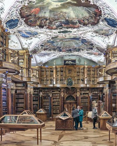 The image shows the interior of a grand, two-story baroque library with ornate ceiling frescoes, wooden bookshelves lining the walls, and visitors viewing display cases on the floor.