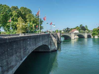 A wide river scene showing a stone arch bridge in the distance under a blue sky with clouds, viewed from a shadowed riverbank with trees and a railing in the foreground.