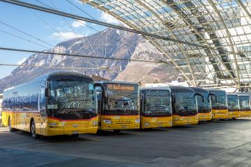 A row of yellow PostAuto buses parked under a large, modern glass and steel canopy structure with mountains visible in the background.