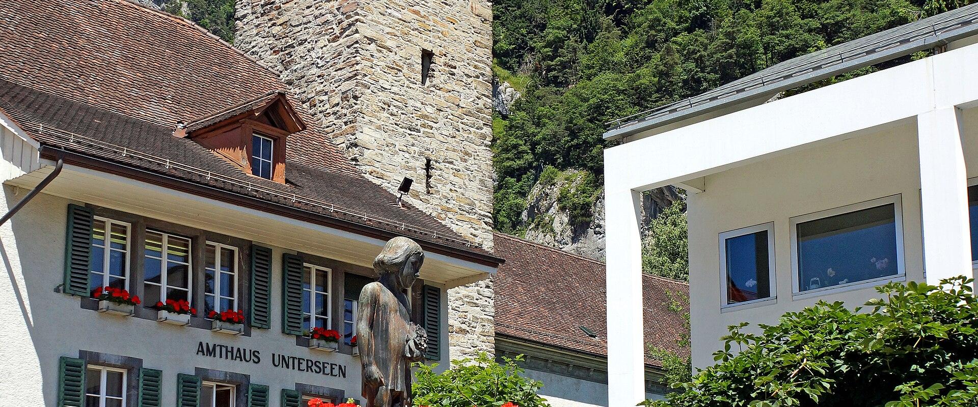 A stone clock tower and a white building labeled 'AMTHAUS UNTERSEEN' with a fountain in the foreground, set against a forested mountain.