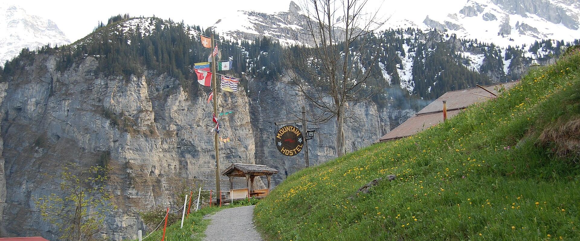 A gravel path leading uphill past a wooden shelter and a sign reading "MOUNTAIN HOSTEL", with a cliff face and snow-capped mountains in the background.