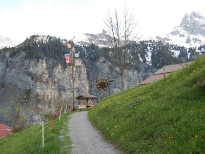 A gravel path leading uphill past a wooden shelter and a sign reading "MOUNTAIN HOSTEL", with a cliff face and snow-capped mountains in the background.