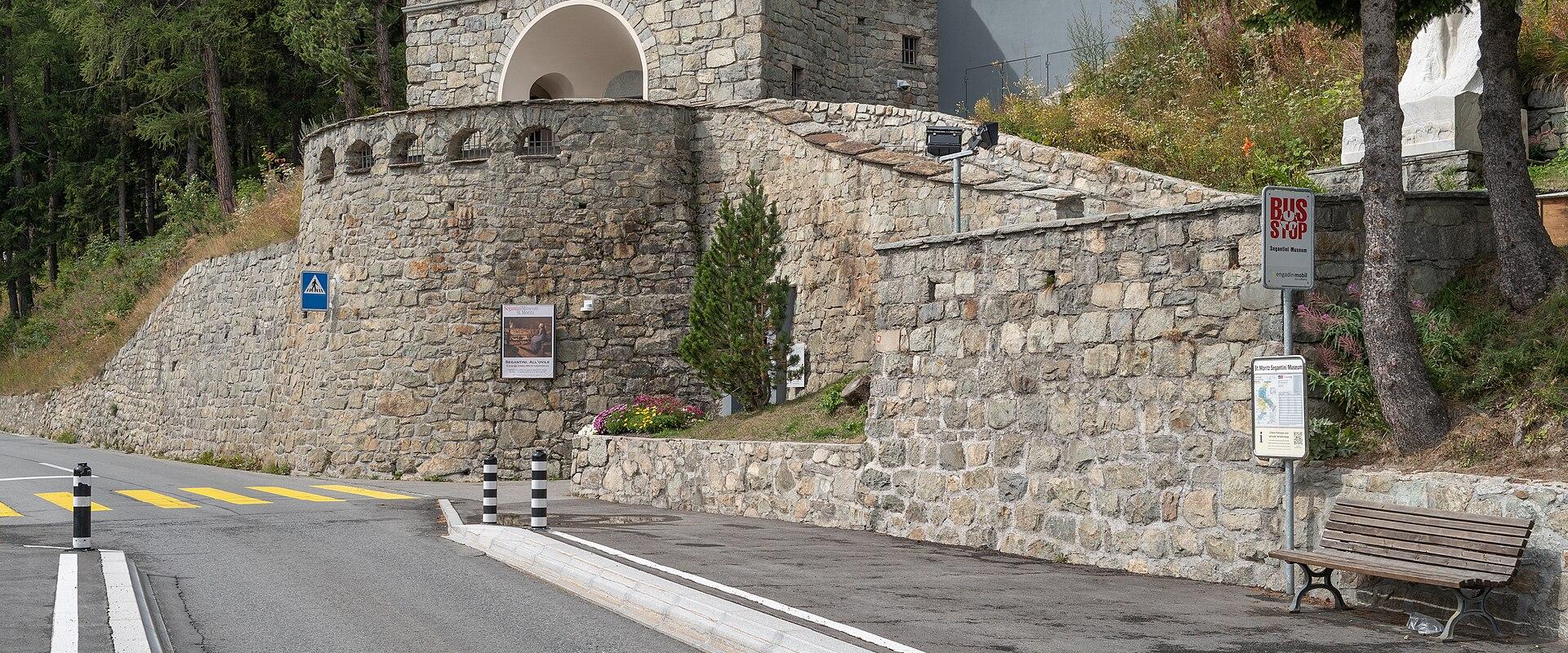 A stone rotunda building with a conical tiled roof situated on a hillside next to a paved road, featuring a bus stop sign and a bench in the foreground.