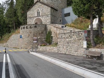 A stone rotunda building with a conical tiled roof situated on a hillside next to a paved road, featuring a bus stop sign and a bench in the foreground.