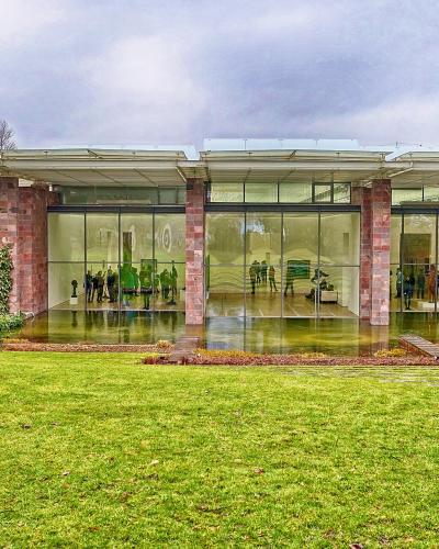 A modern museum building with a glass facade and red sandstone columns, situated in a green park with visitors visible inside.