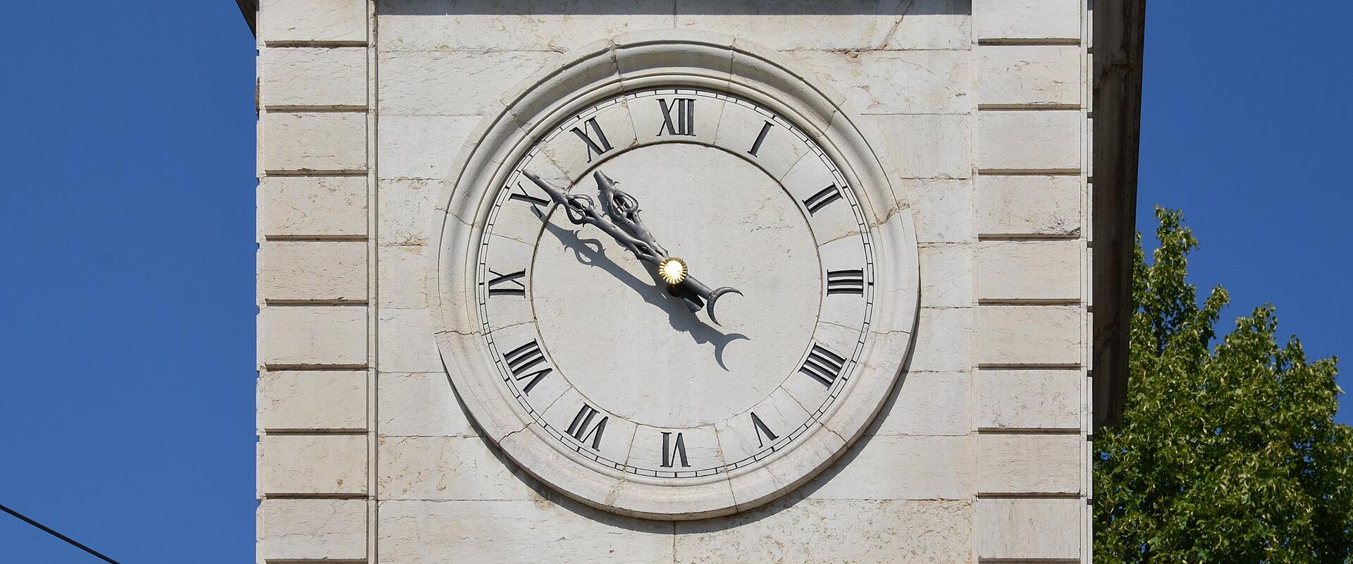 A close-up view of a stone clock tower face with Roman numerals, ornate hands, and a sun/moon indicator against a blue sky.
