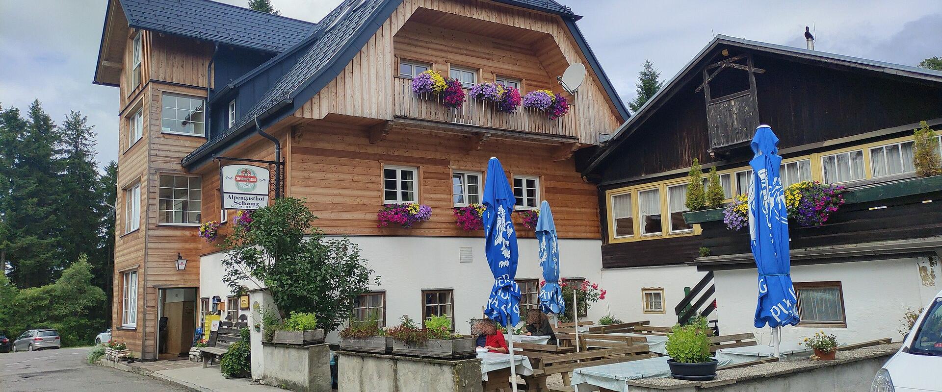 A traditional wooden Alpine inn with flower boxes on the balcony, outdoor seating with blue umbrellas, and a hanging sign reading "Alpenhotel Passhöhe".
