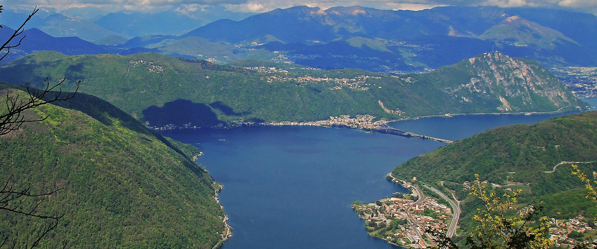 A high-angle scenic view overlooking Lake Lugano, showing the lake's shoreline, surrounding green hills, distant mountains, and a town along the coast, framed by tree branches in the foreground.