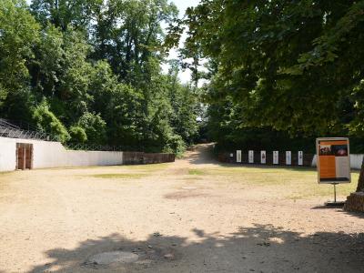 A dirt path leading through a wooded area with low stone walls, informational signs, and a metal railing on the left.