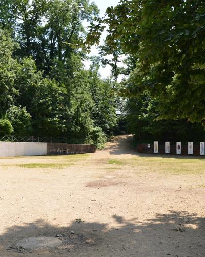 A dirt path leading through a wooded area with low stone walls, informational signs, and a metal railing on the left.