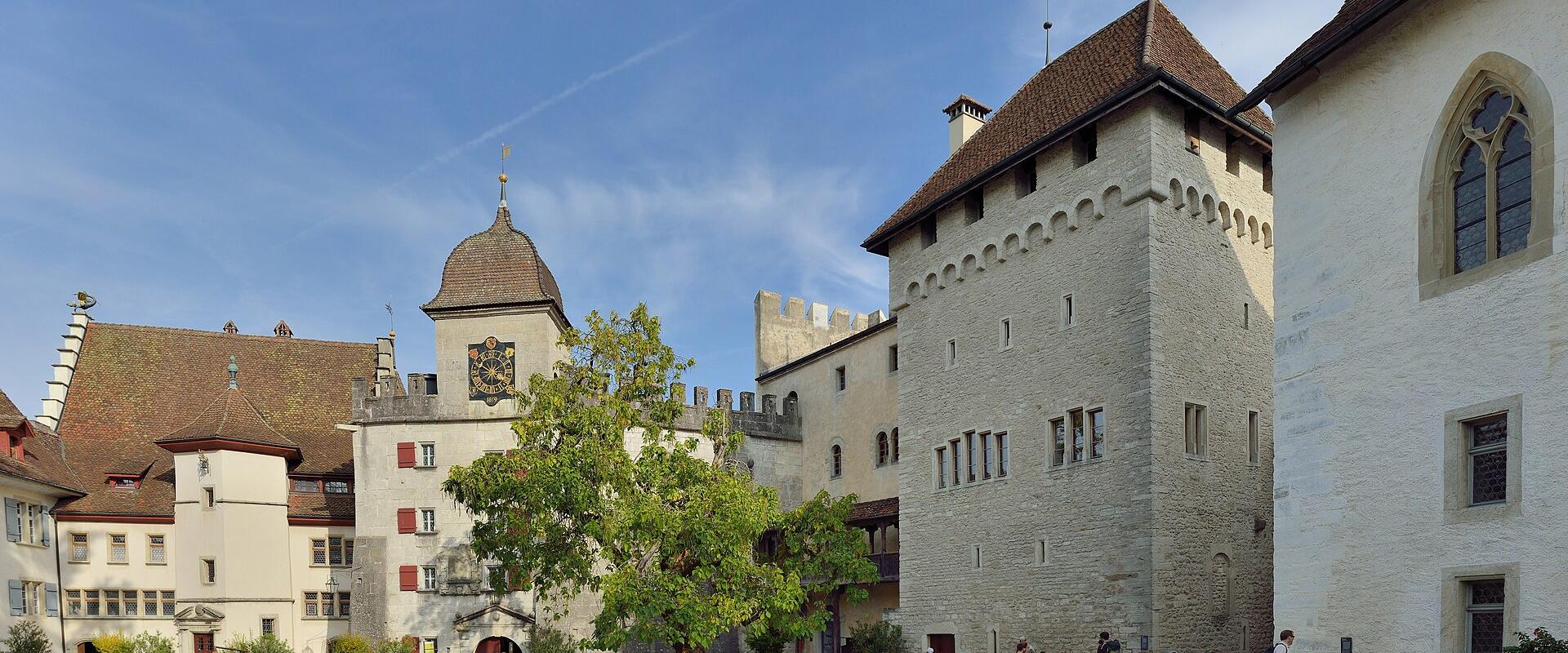 A sunlit courtyard of a medieval castle complex featuring a prominent stone tower with a conical roof, a building with a large coat of arms, and visitors walking on the gravel ground.