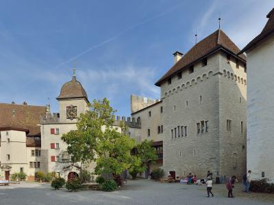 A sunlit courtyard of a medieval castle complex featuring a prominent stone tower with a conical roof, a building with a large coat of arms, and visitors walking on the gravel ground.
