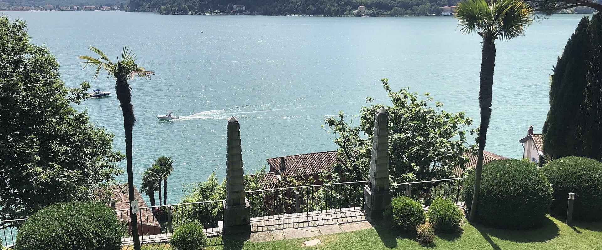 A manicured garden lawn with a stone path, palm trees, and stone obelisks overlooking a large lake with mountains in the background.