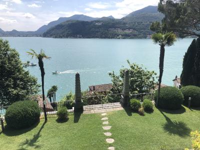 A manicured garden lawn with a stone path, palm trees, and stone obelisks overlooking a large lake with mountains in the background.