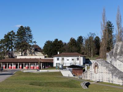 A view of the Roman museum building with its distinctive colonnade and red lower walls, situated next to a white house and the stone ruins of the ancient city walls at Augusta Raurica.