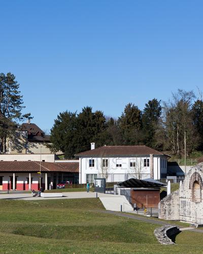 A view of the Roman museum building with its distinctive colonnade and red lower walls, situated next to a white house and the stone ruins of the ancient city walls at Augusta Raurica.