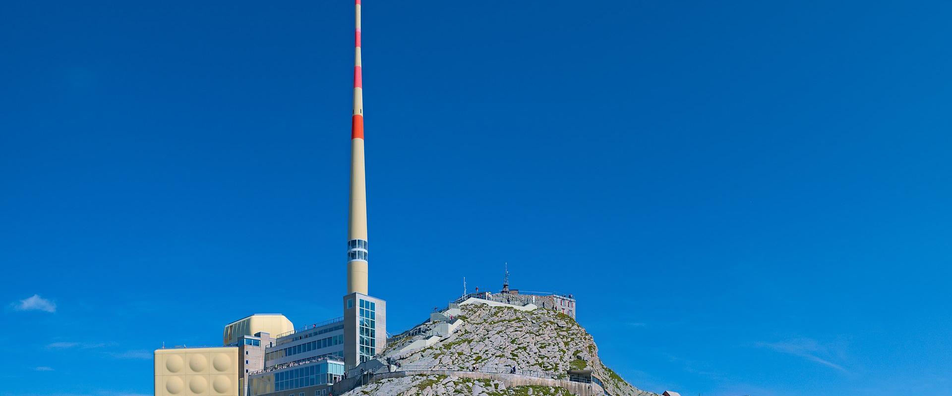 A mountain summit featuring a tall red-and-white striped telecommunications mast, a modern building complex with a distinctive yellow panelled wall, and a traditional red wooden building on a rocky ridge under a clear blue sky.