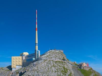 A mountain summit featuring a tall red-and-white striped telecommunications mast, a modern building complex with a distinctive yellow panelled wall, and a traditional red wooden building on a rocky ridge under a clear blue sky.