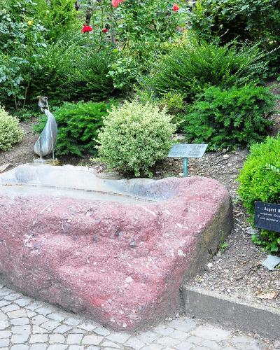 A garden scene featuring a large, rough-hewn pinkish-red stone basin with a small metal sculpture of a bird or figure emerging from it, surrounded by green shrubs and rose bushes, with a black informational sign in the foreground.