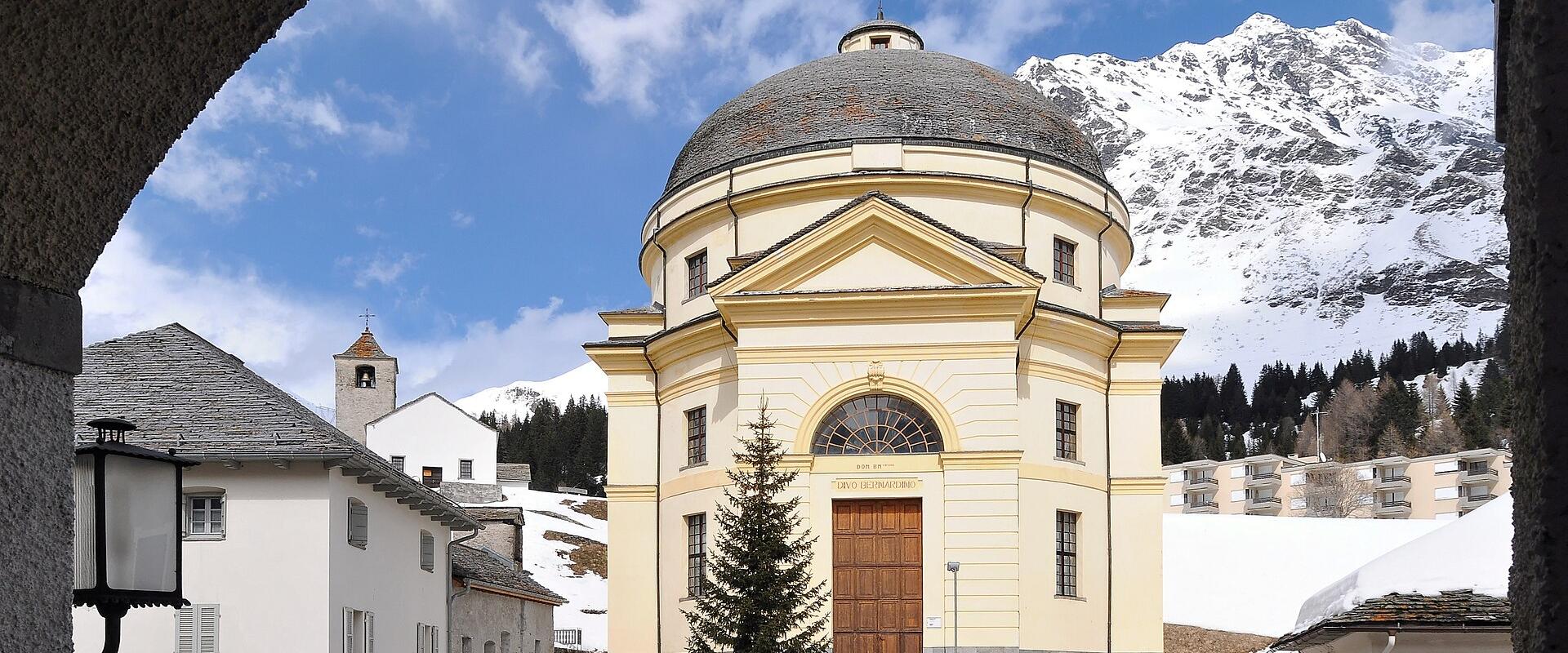 A yellow neoclassical church with a dome and a wooden door, viewed through a stone archway, with snow-capped mountains in the background.