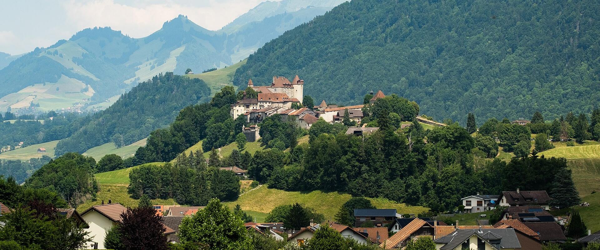 A scenic landscape view of a medieval castle and fortified village situated on a forested hill, with modern houses in the foreground and mountains in the background.