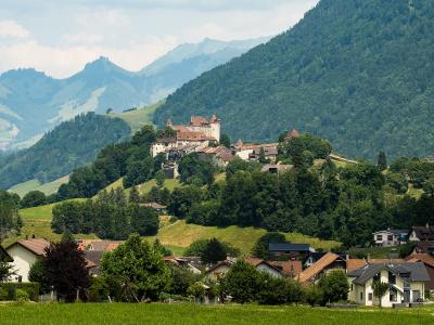 A scenic landscape view of a medieval castle and fortified village situated on a forested hill, with modern houses in the foreground and mountains in the background.