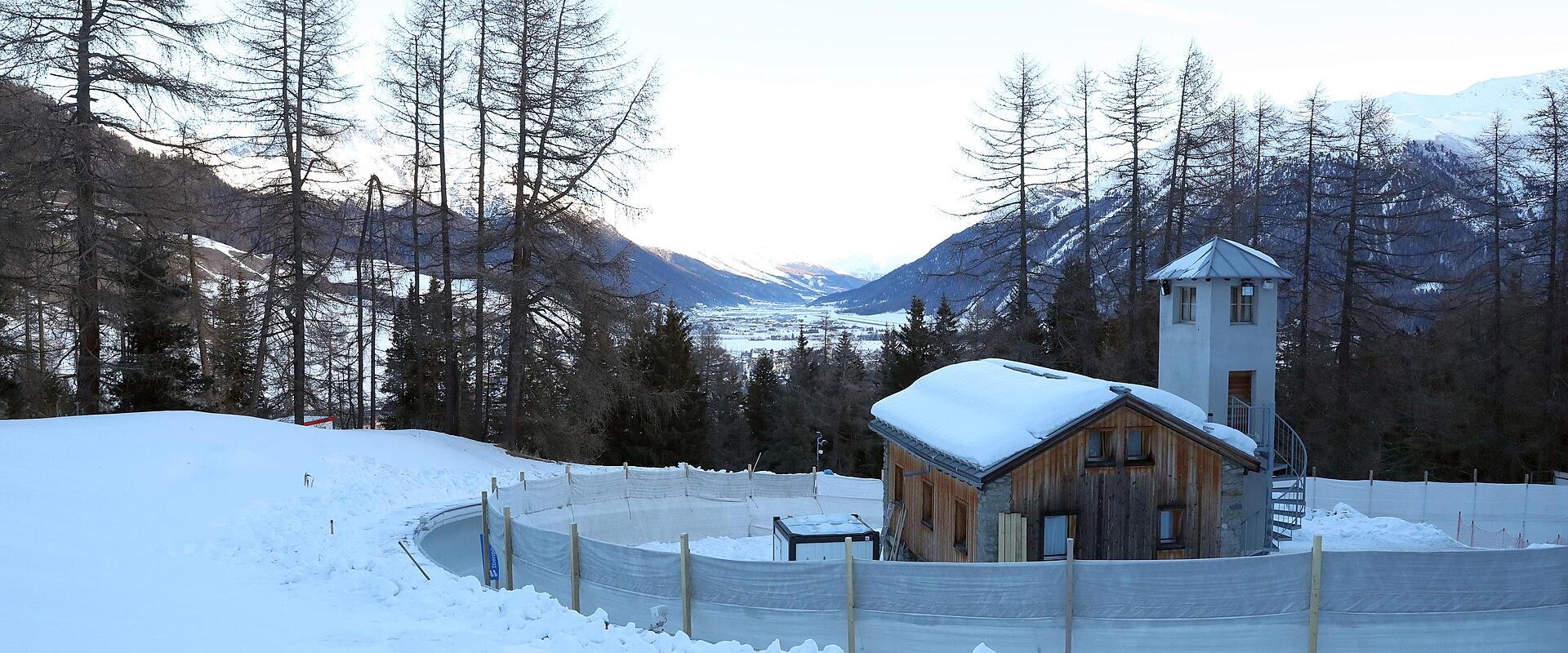 A snowy bobsleigh track curve with safety netting, a wooden building, and a watchtower, set against a backdrop of mountains and trees.