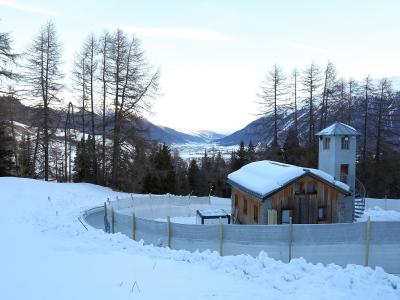 A snowy bobsleigh track curve with safety netting, a wooden building, and a watchtower, set against a backdrop of mountains and trees.