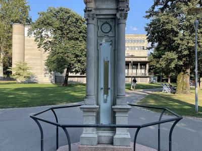 A stone weather station monument featuring a barometer and clock, topped with a weather vane, enclosed by a low metal railing on a paved path in a park.
