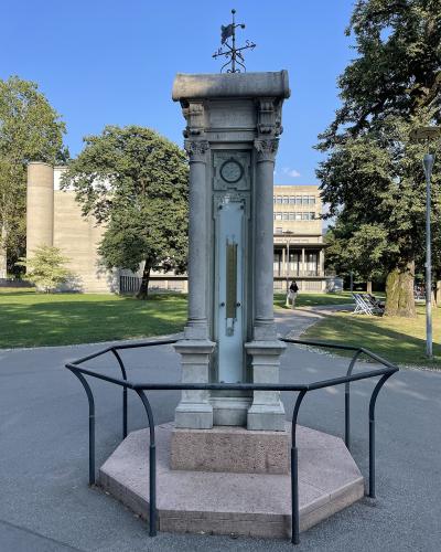 A stone weather station monument featuring a barometer and clock, topped with a weather vane, enclosed by a low metal railing on a paved path in a park.