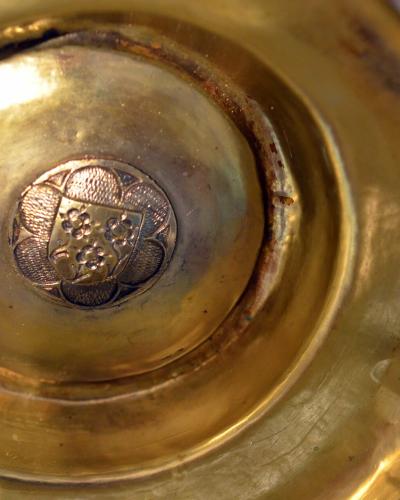 A close-up photograph of the interior bottom of a golden metal drinking vessel, featuring an embossed coat of arms in the center and an ornate handle attachment visible on the right edge.
