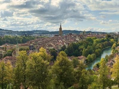 A panoramic view of a European city with a prominent cathedral spire, a river curving through the center, and multiple bridges, taken from an elevated park-like vantage point.