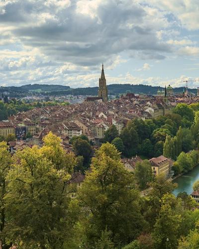 A panoramic view of a European city with a prominent cathedral spire, a river curving through the center, and multiple bridges, taken from an elevated park-like vantage point.