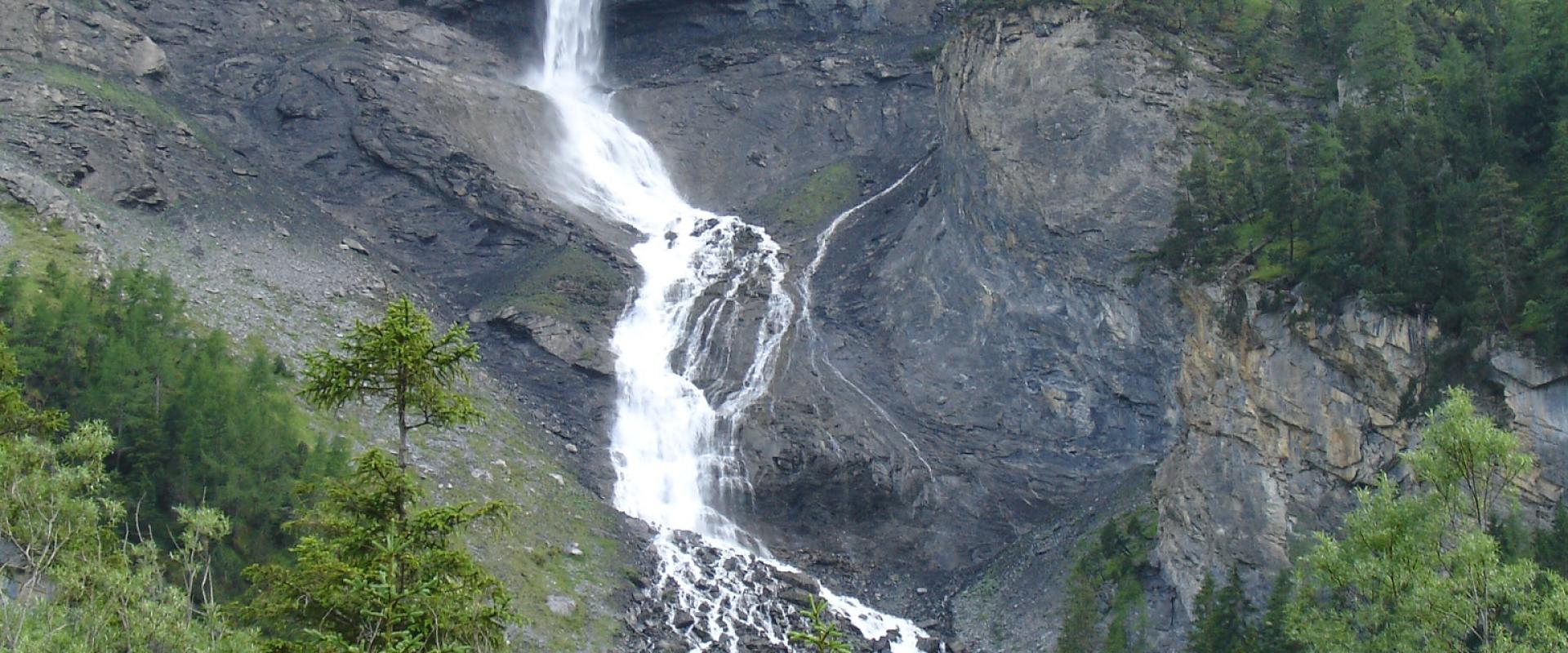 A tall, single-stream waterfall cascading down a steep, grey rock face into a rocky basin, surrounded by green coniferous trees in the foreground.