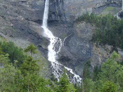 A tall, single-stream waterfall cascading down a steep, grey rock face into a rocky basin, surrounded by green coniferous trees in the foreground.