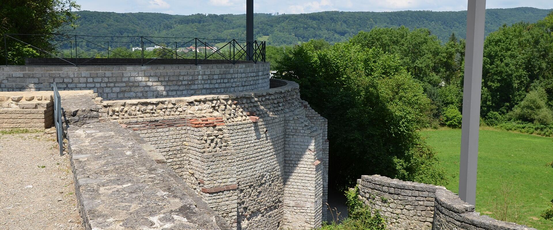 The image shows a section of reconstructed Roman stone walls and foundations, featuring brickwork and stone masonry, situated within a grassy archaeological site with a modern viewing platform structure overhead.