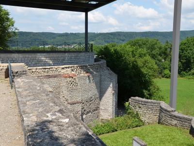 The image shows a section of reconstructed Roman stone walls and foundations, featuring brickwork and stone masonry, situated within a grassy archaeological site with a modern viewing platform structure overhead.
