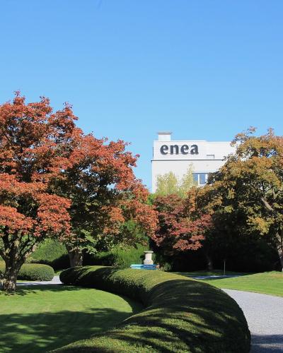 A landscaped park with manicured lawns, gravel paths, sculpted hedges, and trees showing autumn foliage, with a white building in the background displaying the word "enea" in large letters.