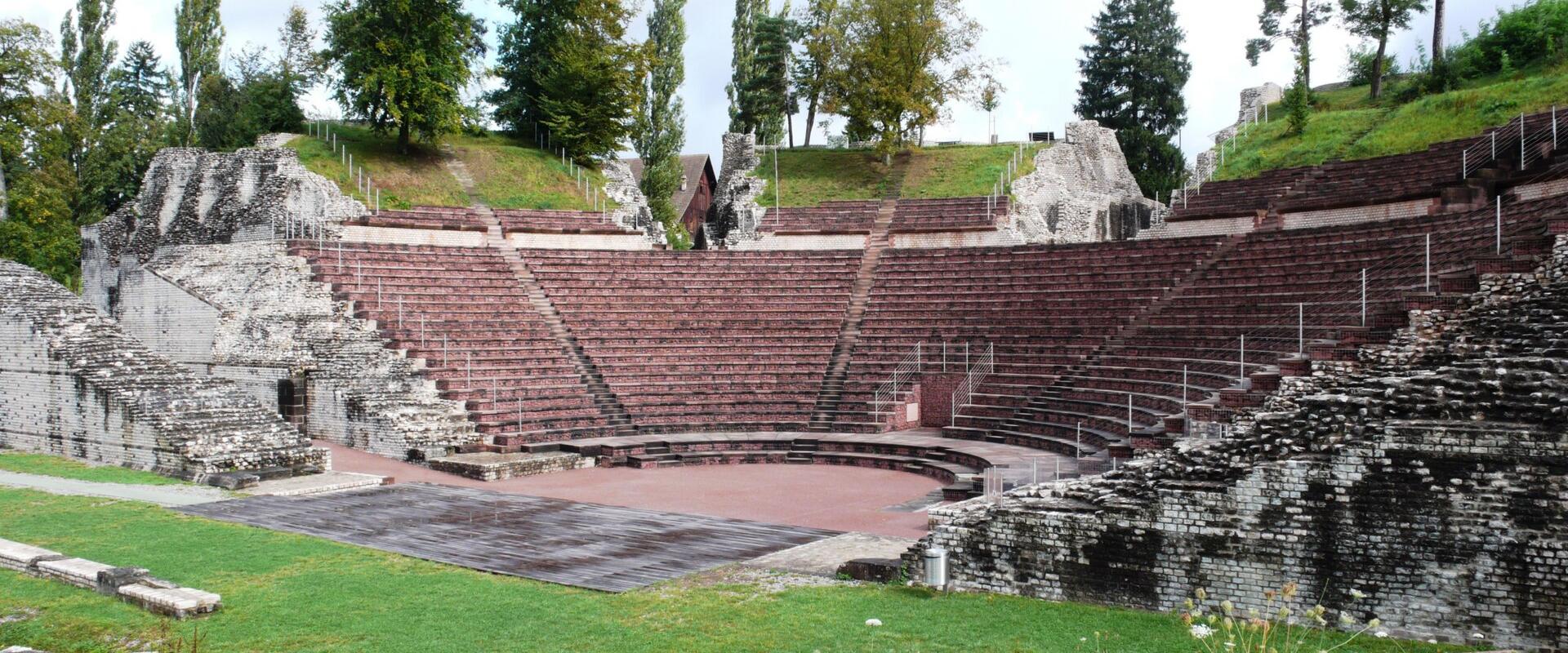 The image shows the ruins of a Roman amphitheater with tiered seating, stone walls, and a central arena floor surrounded by grass and trees.