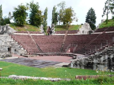 The image shows the ruins of a Roman amphitheater with tiered seating, stone walls, and a central arena floor surrounded by grass and trees.