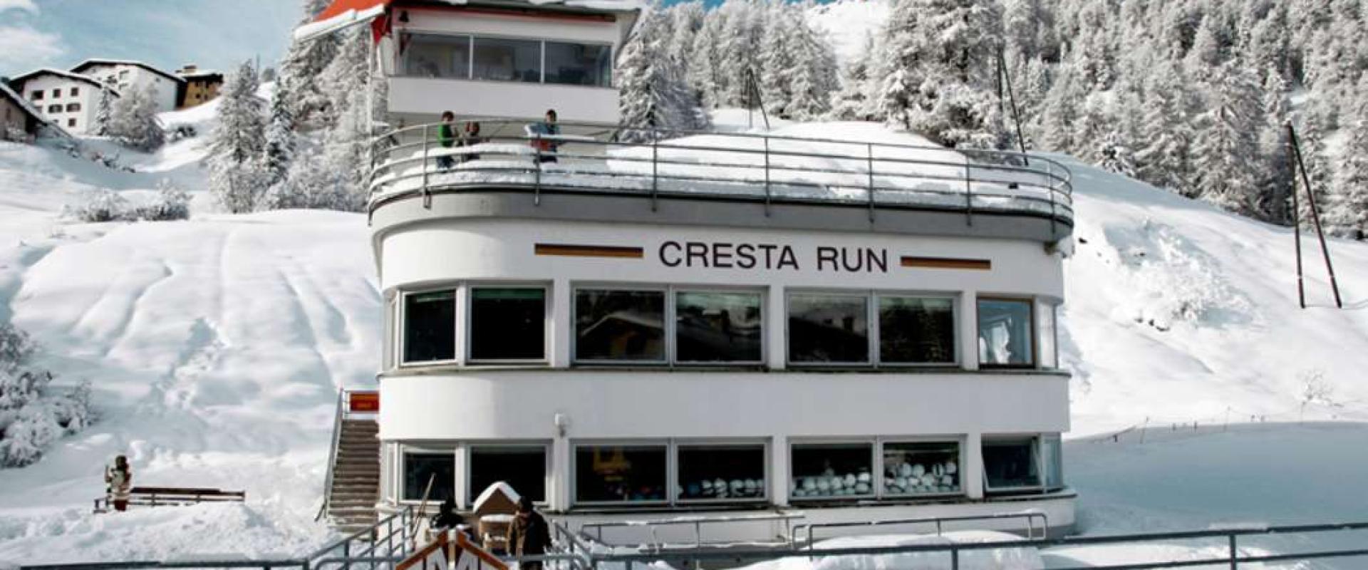 A white, multi-story building with a curved facade situated in a snowy alpine landscape, featuring large windows and a balcony with people standing on it.