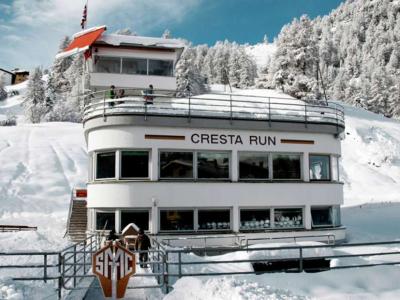 A white, multi-story building with a curved facade situated in a snowy alpine landscape, featuring large windows and a balcony with people standing on it.